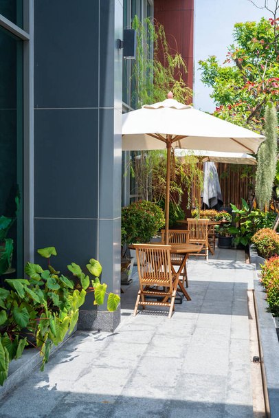 Ξύλινες καρέκλες και τραπεζάκια με ομπρέλες στο μπαλκόνι του ξενοδοχείου με πράσινη διακόσμηση φυτών σε ηλιόλουστη μέρα. Κάθεστε να πιείτε ζεστό καφέ ή να πάρετε τσιγάρα. - Φωτογραφία, εικόνα
