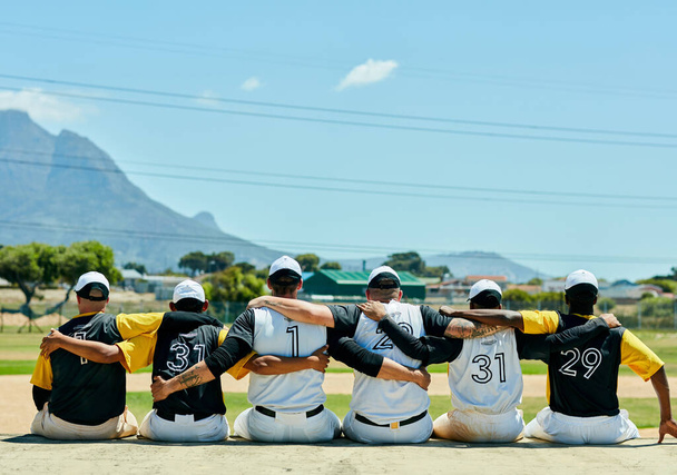 Єдність завжди перемагає. Знімок заднього виду команди невпізнаваних бейсболістів, які обіймають один одного, сидячи біля бейсбольного поля протягом дня
 - Фото, зображення