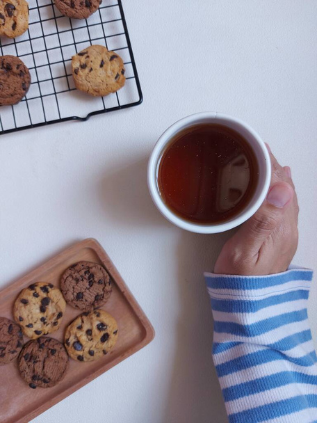 Tea time photography concept.  A cup of tea. Completed with choco chips cookies, white napkin and cooling rack. Isolated background in white. Minimalist and aesthetic photo. Hand in frame - Photo, Image