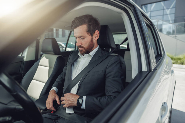 Όμορφος επιχειρηματίας δένει τη ζώνη του καθισμένος στο αυτοκίνητο. Θολή φόντο - Φωτογραφία, εικόνα