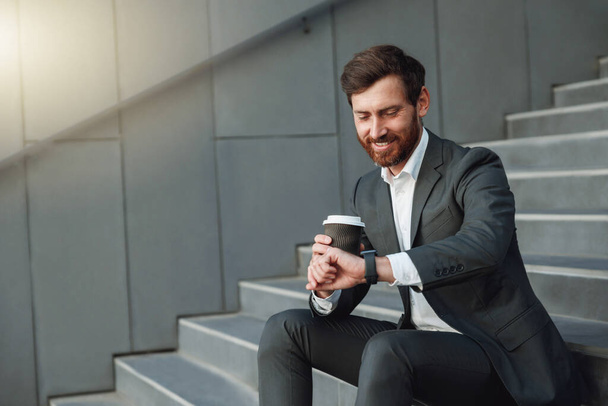 Businessman in suit sitting on stairs with coffee during break. Blurred background - Foto, imagen