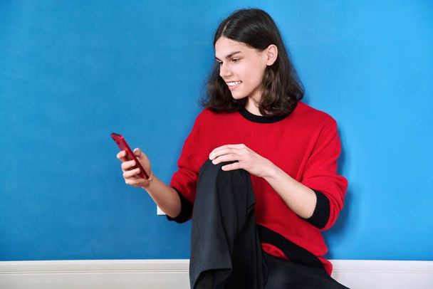 Teenage trendy male with a smartphone in his hands sitting on the floor on a blue background. Youth, lifestyle, communication, leisure, color, young people concept - Fotó, kép