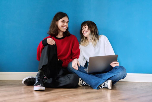 Couple of laughing cheerful teenage students using laptop sitting on the floor on blue background. E-education, online learning, youth, friendship, lifestyle, leisure, technology concept - Foto, afbeelding