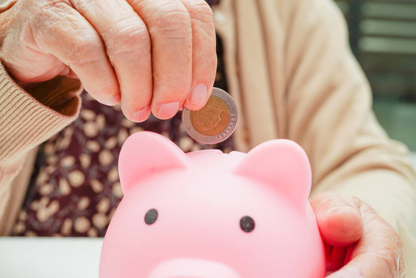 Retired elderly woman putting coins money in piggy bank and worry about monthly expenses and treatment fee payment. - Photo, image