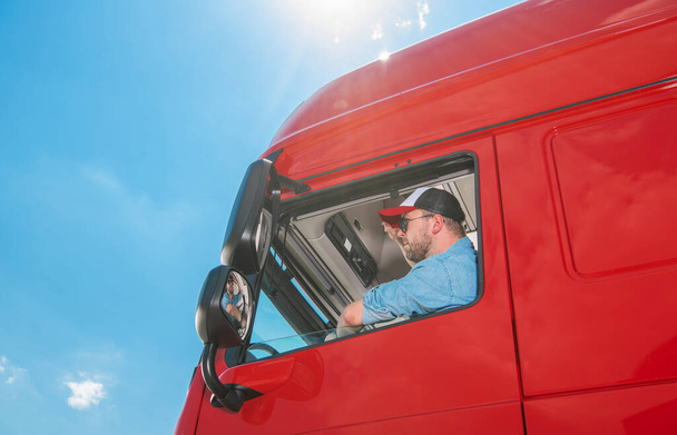 Truck Driver in a Blue Shirt and Baseball Cap Sitting in His Huge Reg Lorry Looking at the Road Ahead. Blue Sunny Sky in the Background. Transportation Theme. - Photo, image