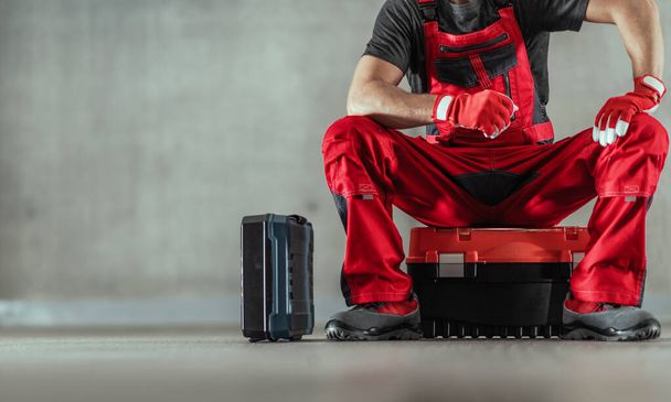 Closeup of Handyman in Red Functional Work Wear and Protective Gloves Sitting on One of His Toolboxes While Waiting for the Client in the Indoor Facility. Solid Grey Background. - Photo, image