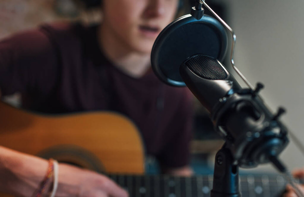 Microphone with pop filter closeup photo with a young teenager boy in headphones recording voice and guitar playing music. Modern home sound studio audio recording technology and teens hobby concept. - 写真・画像