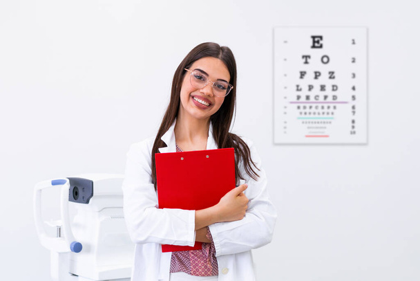Улыбающийся офтальмолог держит планшет на фоне диаграммы глаз. Докторша-окулистка с глазной картой за спиной - Фото, изображение