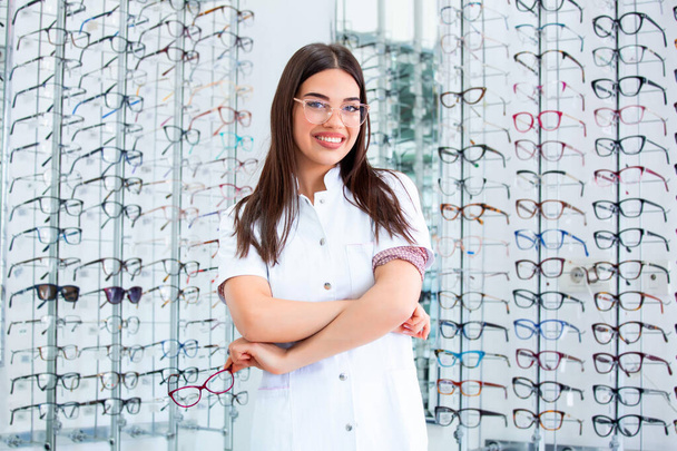Jovem médica atraente na clínica de oftalmologia. Médico oftalmologista está de pé perto prateleiras com óculos diferentes
 - Foto, Imagem