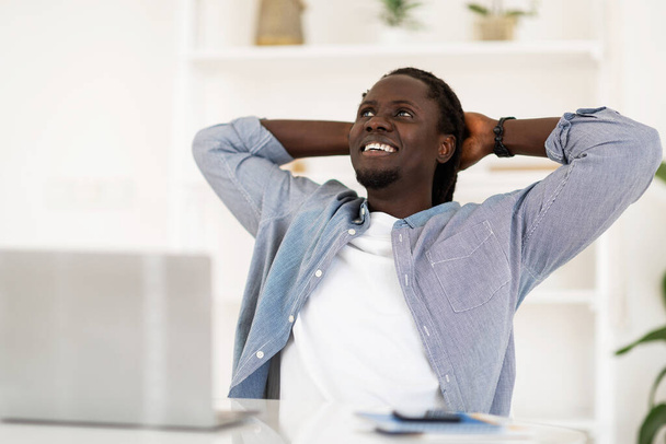 Relaxed Black Male Freelancer Leaning Back In Chair, Resting After Online Work With Laptop In Home Office, Dreamy Young African American Man Sitting At Desk With Hands Behind Head And Looking Aside - Photo, image