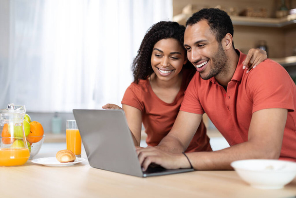 Glad young african american wife and husband have breakfast in morning together, enjoy chat, watch video and shopping online on laptop in kitchen interior. Video call, app and food blog, new normal - Foto, immagini