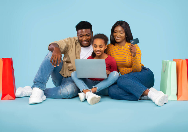 Happy black family shopping online with laptop and credit card, making payment while sitting on floor over blue background. Parents and daughter buying something in internet - Valokuva, kuva