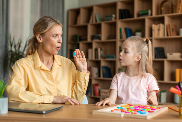 Caring mother teaching small preschool child daughter pronouncing words and letters correctly. Smart girl doing vocal exercises with professional teacher, sitting at desk - Foto, Imagen