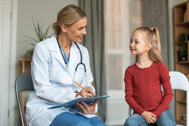 Pleasant female pediatrician talking to pretty schoolgirl patient and writing down notes in medical history paper during home calling visit. Healthcare concept - Foto, afbeelding