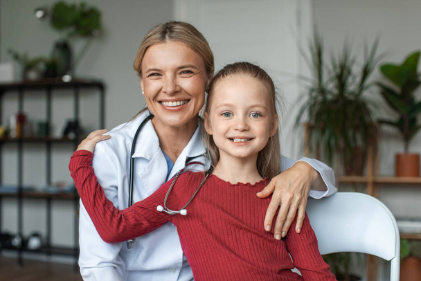 Πορτρέτο του φιλικό νοσοκόμα σε λευκό παλτό, αγκαλιάζει με ευτυχισμένη μικρή ασθενή προσχολικής ηλικίας, χαμογελαστό κορίτσι ποζάρουν για φωτογραφία με ελκυστικό γενικό ιατρό - Φωτογραφία, εικόνα