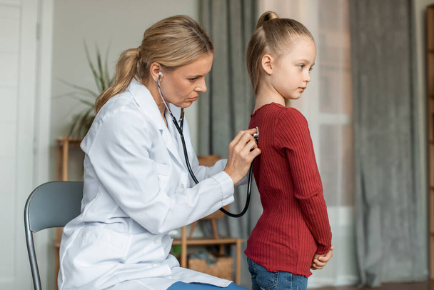 Female pediatrician listening to heartbeat and lungs by applying stethoscope to back of little girl. Doctor came to home to examine the child - Foto, imagen