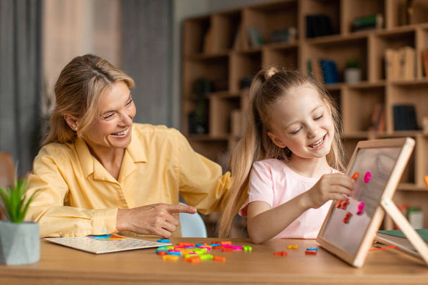 Счастливая учительница и маленькая девочка учатся, ребенок делает работы на флип-чарте, сидя за партой в офисе. Положительный частный репетитор, занимающийся с учеником. Концепция образования - Фото, изображение
