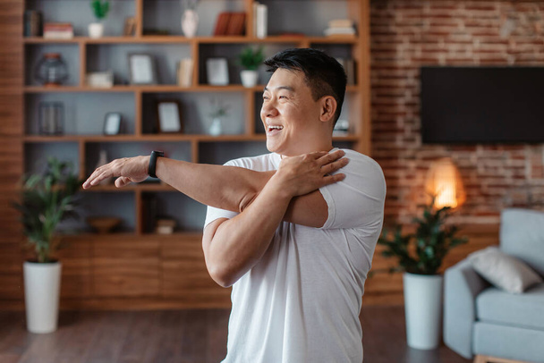Active asian mature man stretching arm muscles, doing warmup exercises at home, working out in living room interior. Middle aged korean male having domestic training - Foto, Imagem