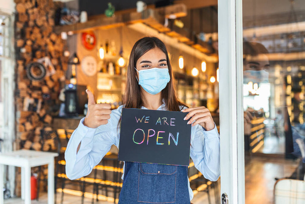 Small business owner with face mask holding the sign for the reopening of the place after the quarantine due to covid-19. Woman with protective mask holding sign we are open, support local business. - Zdjęcie, obraz