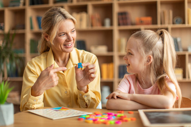 Happy woman speech therapist teaching little girl with pronounciation deffects to say sound R during personal training at classroom - Photo, image
