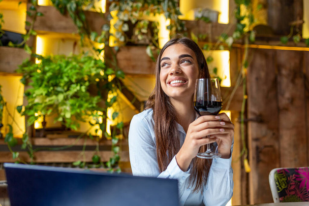 Η γυναίκα δουλεύει με ένα ποτήρι κόκκινο κρασί και λάπτοπ στο καφέ. Γυναίκα που χρησιμοποιεί φορητό υπολογιστή ενώ έχει κρασί στο εστιατόριο. ελεύθερος επαγγελματίας - Φωτογραφία, εικόνα