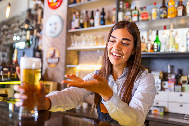 Красивая улыбающаяся барменша, подающая пиво за стойкой бара, полки полные бутылок с алкоголем на заднем плане - Фото, изображение