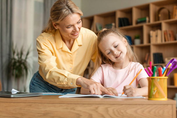 Caring mother helping to adorable little daughter with homework, sitting at table in living room, smart girl doing school tasks, studying, homeschooling - Zdjęcie, obraz