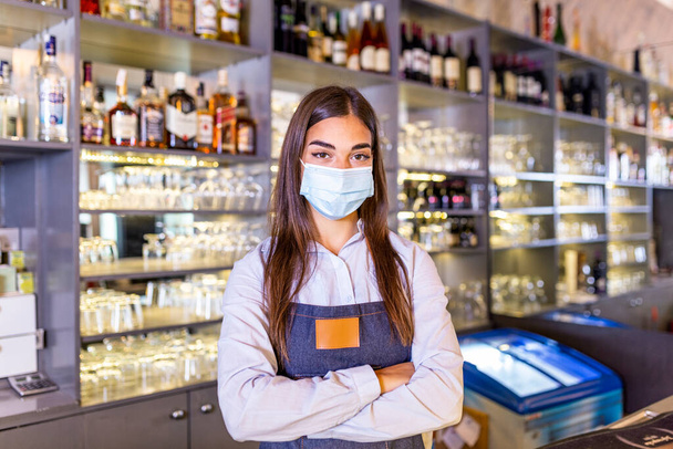 Portrait of a beautiful bartender standing at the counter smiling and looking at the camera while while wearing protective face mask due to covid-19, shelves full of bottles with alcohol - Fotografie, Obrázek