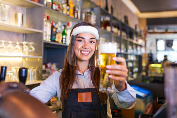 Bartender with face shield,covid-19 protection, serving a draft beer at the bar counter during coronavirus pandemic, showing OK sign,shelves full of bottles with alcohol on the background - Foto, Imagen
