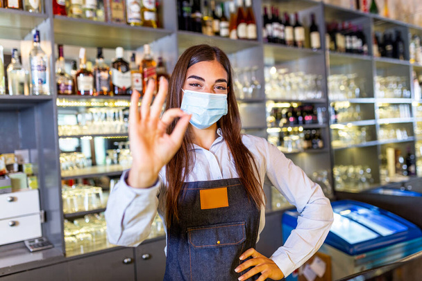Happy young woman wearing protective face mask in the retaurant during coronavirus pandemic, Bartender showing Ok sign. Shelves full of bottles with alcohol on the background - Valokuva, kuva