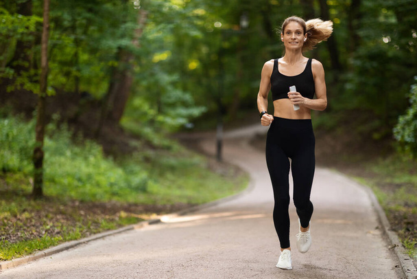 Joyful attractive athletic woman jogging by public park, holding smartphone, listening to music while exercising, getting ready for marathon, panorama with copy space, full length - Foto, imagen