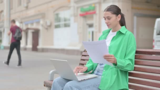 Νεαρή γυναίκα ανάγνωση εγγράφων και χρήση στο φορητό υπολογιστή, ενώ κάθεται σε εξωτερικούς χώρους - Πλάνα, βίντεο