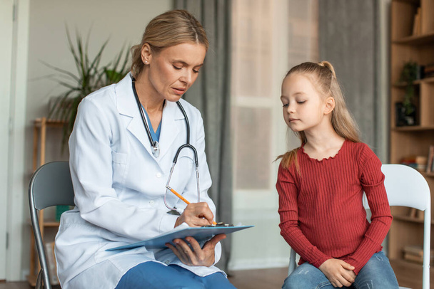 Focused female doctor writing down girls symptoms on clipboard while child looking what doc writing in chart, sitting next to her - Valokuva, kuva