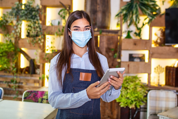 Portrait of beautiful waitress wearing protective face mask while holding touchpad and looking at the camera in a pub or restaurant during COVID-19 epidemic. - Photo, image