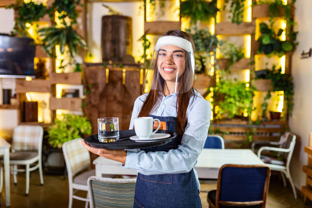 Female waitress wearing face shield, visor serves the coffee in restaurant during coronavirus pandemic representing new normal concept - Fotó, kép