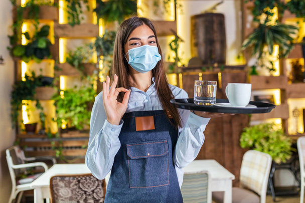 Γυναίκα σερβιτόρα σε μια ιατρική προστατευτική μάσκα σερβίρει τον καφέ στο εστιατόριο durin coronavirus πανδημία αντιπροσωπεύει νέα κανονική έννοια και δείχνει OK σημάδι - Φωτογραφία, εικόνα