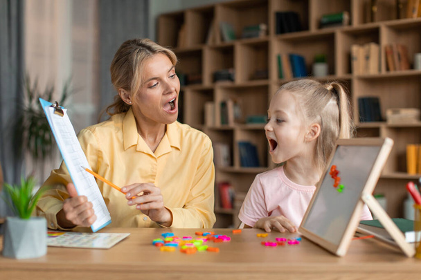 Speech training for kids. Professional female specialist training with little girl at cabinet, teaching her right articulation exercises, showing something on clipboard - Foto, imagen