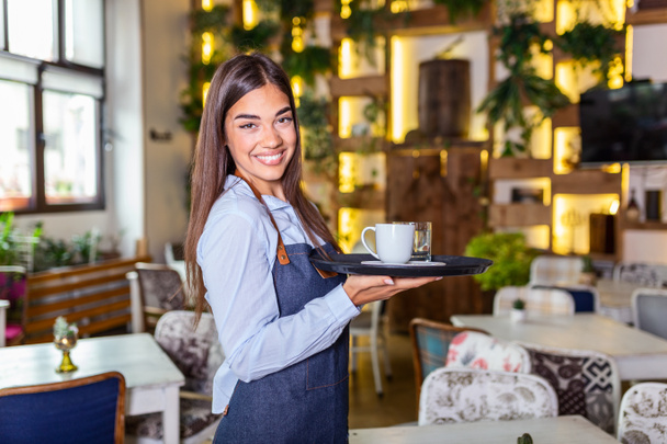 Счастливая официантка держит поднос с чашкой кофе, работает в кафетерии и обслуживает стол - Фото, изображение