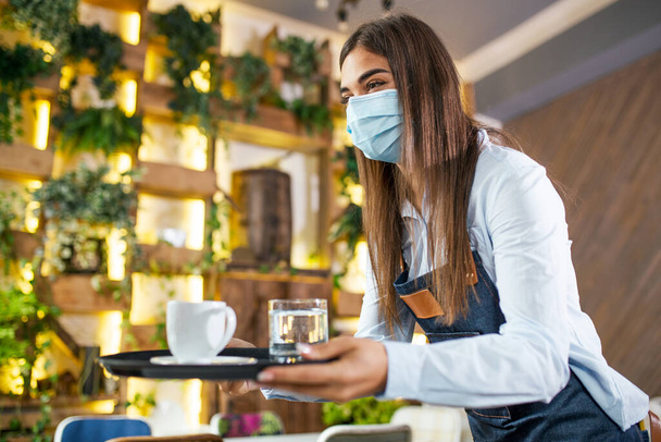 Waitress serving drinks in restaurant or cafe. Young woman wearing protective face mask and serving drinks during coronavirus pandemic - Photo, image