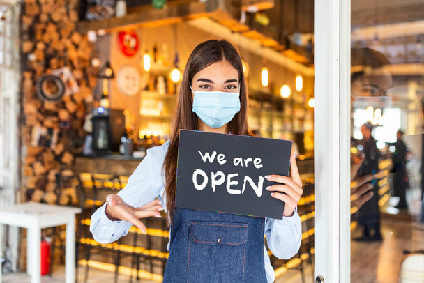 Small business owner with face mask holding the sign for the reopening of the place after the quarantine due to covid-19. Woman with protective mask holding sign we are open, support local business. - Fotoğraf, Görsel