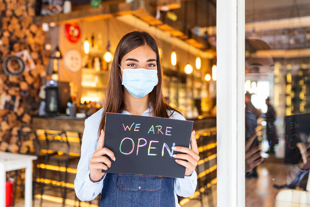 Small business owner with face mask holding the sign for the reopening of the place after the quarantine due to covid-19. Woman with protective mask holding sign we are open, support local business. - Photo, image