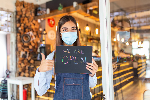 Happy female waitress with protective face mask holding open sign while standing at cafe or restaurant doorway, open again after lock down due to outbreak of coronavirus covid-19. Showing OK sign - Foto, afbeelding