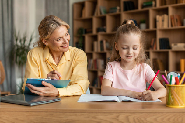 Happy female tutor sitting at table with smart little girl, helping child with homework, preparing school assignments together at home. Homeschooling concept. - Foto, Bild