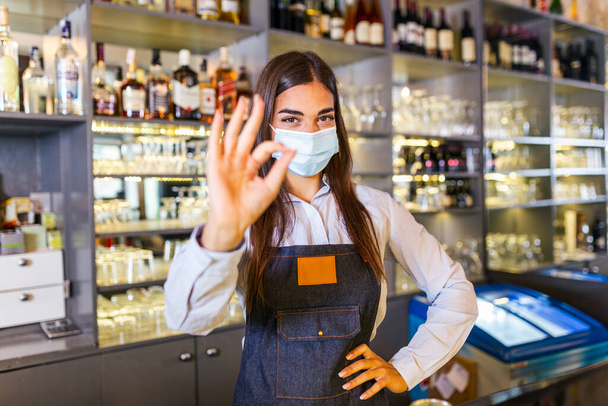 Щаслива молода жінка, одягнена в захисну маску під час пандемії коронавірусу, Бартендер показує знак Ок. Залишки пляшок з алкоголем на задньому плані - Фото, зображення