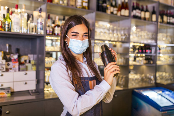 Νεαρή εργάτρια σε ένα μπαρ που ετοιμάζει κοκτέηλ με σέικερ. Όμορφη νεαρή γυναίκα πίσω από το μπαρ φορώντας προστατευτική μάσκα προσώπου - Φωτογραφία, εικόνα