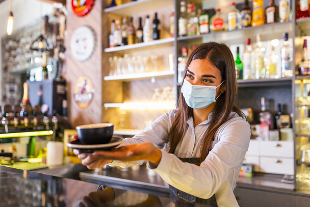 Beautiful female barista is holding a cup with hot coffee, looking at camera and wearing protective face mask while standing near the bar counter in cafe - Photo, image