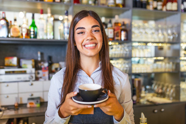 Красива бариста тримає чашку з гарячою кавою, дивлячись на камеру і посміхаючись, стоячи біля барної стійки в кафе
 - Фото, зображення