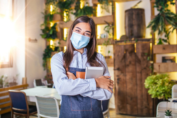 Happy female waitress using digital tablet while wearing protective face mask at the restaurant or cafe. Open again after lock down due to outbreak of coronavirus covid-19, New normal - Foto, imagen