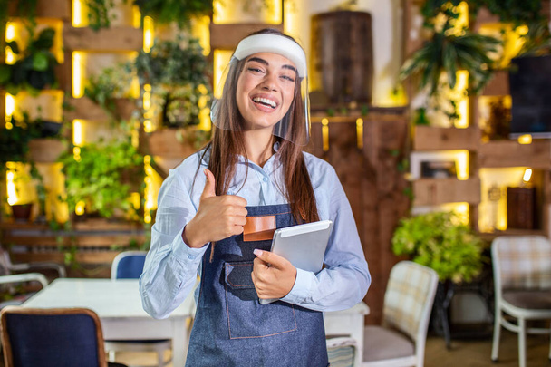 Happy female waitress using digital tablet while wearing visor at the restaurant or cafe. Open again after lock down due to outbreak of coronavirus covid-19, New normal - Zdjęcie, obraz