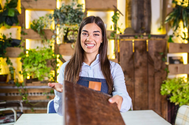 Happy beautiful smiling waitress wearing apron giving a folder menu in a restaurant, looking at camera, standing in cozy coffeehouse, good service - Photo, Image